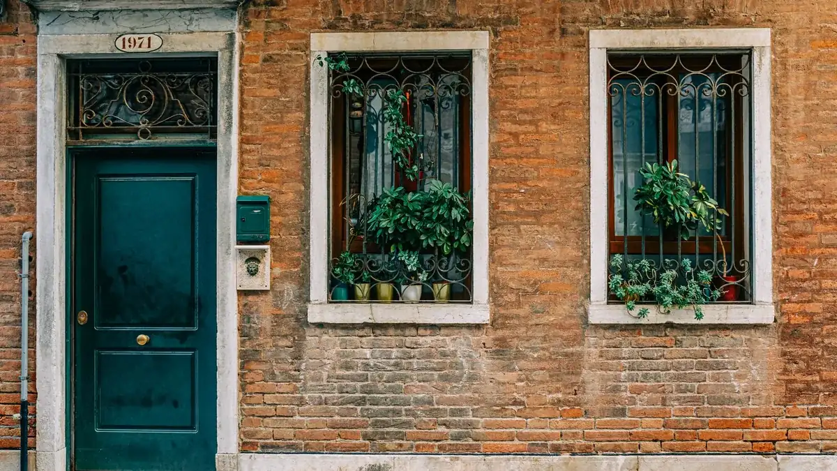 Fachada antigua de ladrillo con dos ventanas de rejería y macetas con plantas, junto a una puerta verde oscuro con el número 1971.