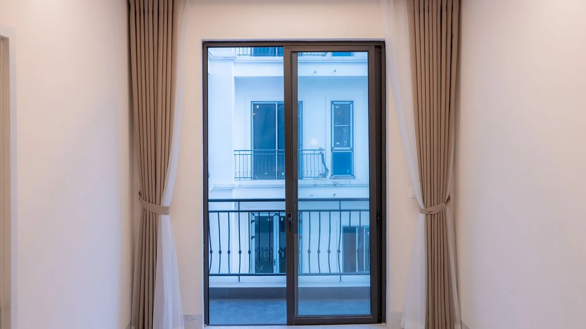 Habitación con una puerta corredera de cristal y marco oscuro que da a un balcón con barandilla de forja, flanqueada por cortinas beige.