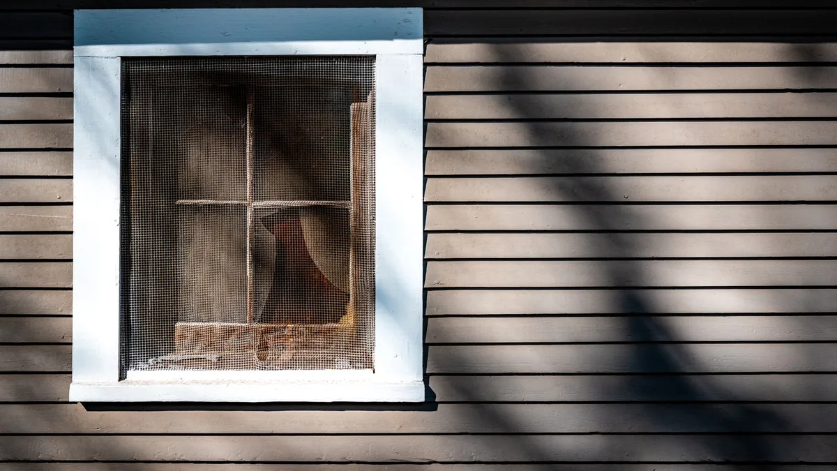 Ventana de marco blanco sobre una fachada de tablillas marrones, con una mosquitera de malla instalada y sombras de ramas proyectadas encima.