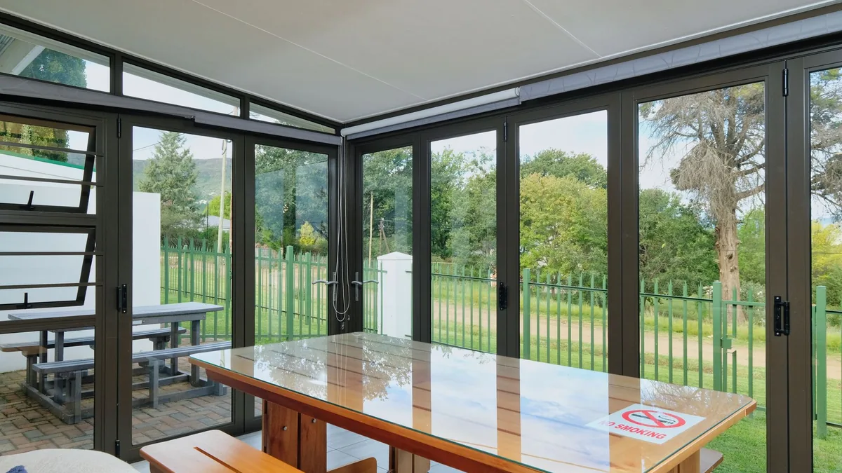 Porche acristalado luminoso con mesa y bancos de madera, y vistas a un jardín vallado a través de las cristaleras plegables de aluminio.