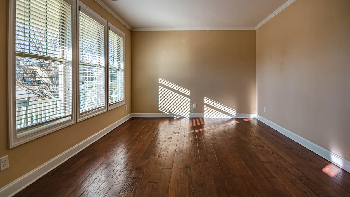 Habitación vacía y luminosa con suelo de madera oscura y tres ventanas de guillotina con persiana, por las que entra la luz proyectando sombras alargadas en el suelo.