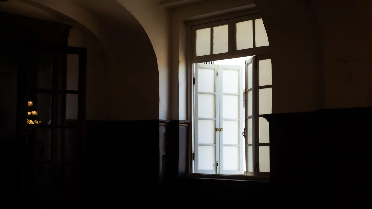 Interior en penumbra de un edificio antiguo, con una ventana de dos hojas y marco blanco abierta de par en par hacia la claridad exterior.