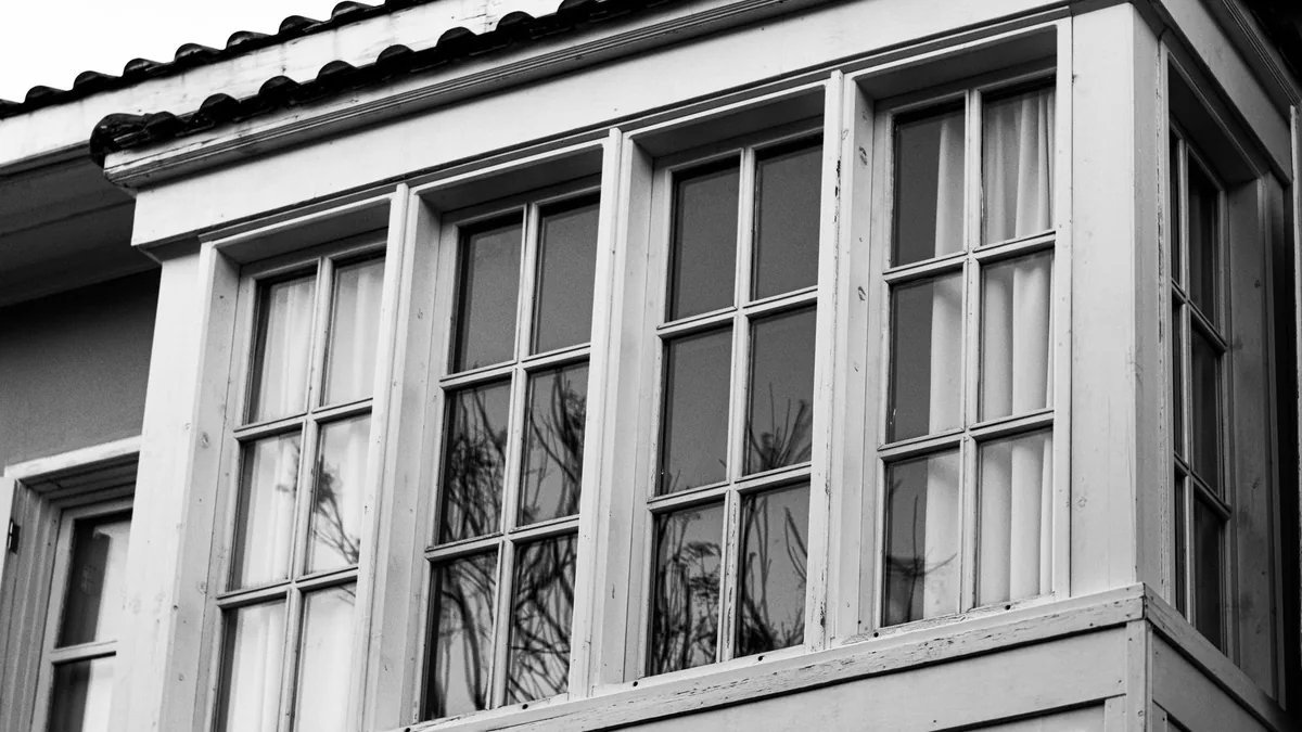 Fotografía en blanco y negro de un mirador con varias hojas de vidrio enmarcadas en madera blanca, reflejando las ramas de un árbol cercano.